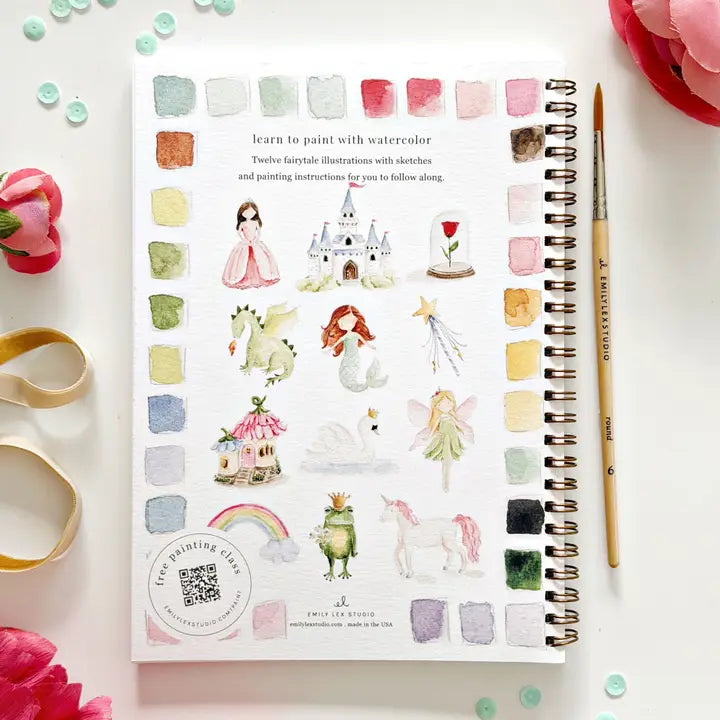Watercolor art book with illustrations of fairies, castles, and other fantasy elements on a white surface with flowers and a paint brush.