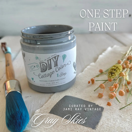 Open 8 oz. Jar of DIY cottage color grey skies, on tan surface, with flowers, brushes, and a swatch surrounding it.