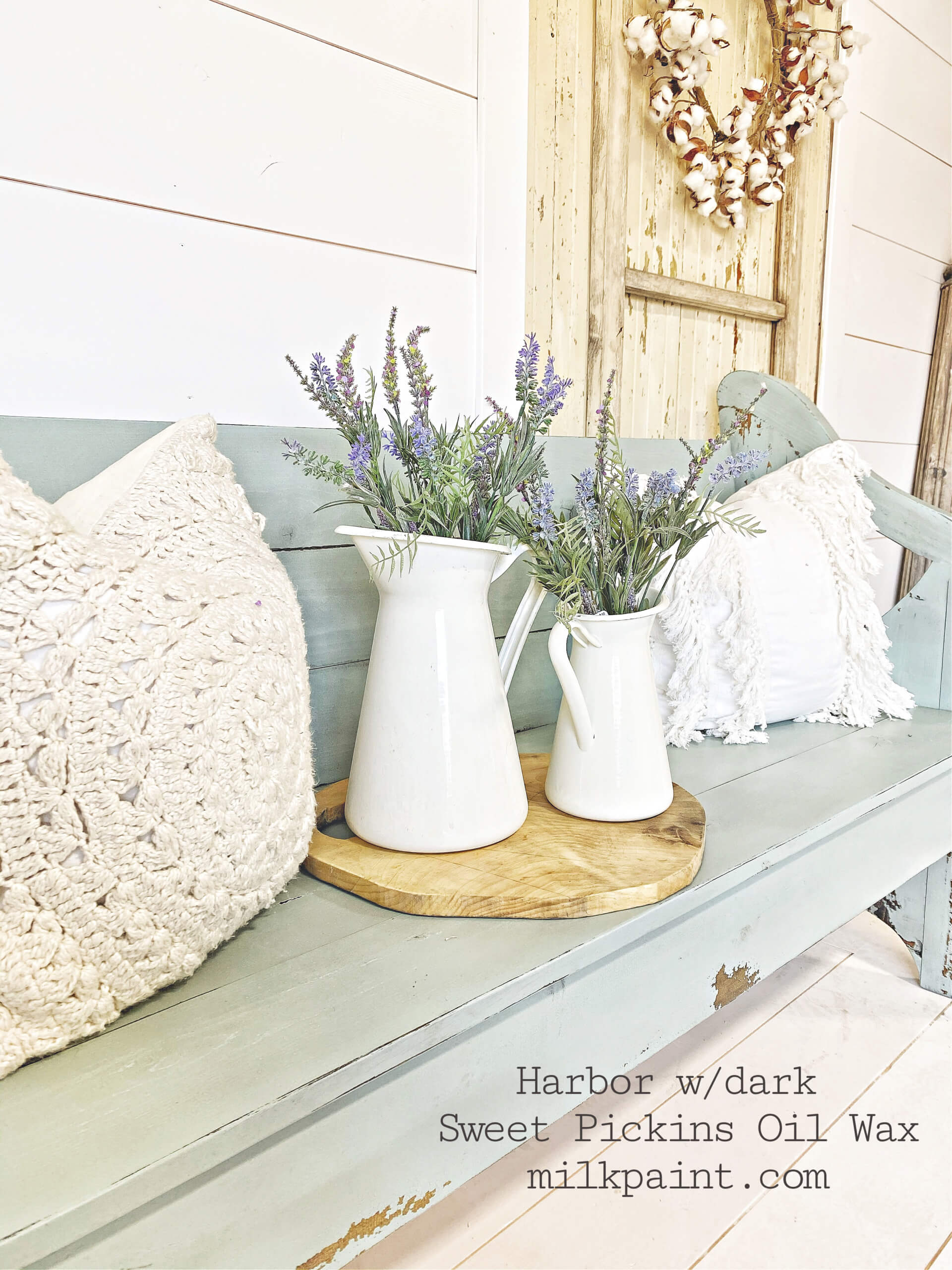 Decorative bench with white pitcher and lavender, surrounded by pillows and a wooden board, on a light wood floor.