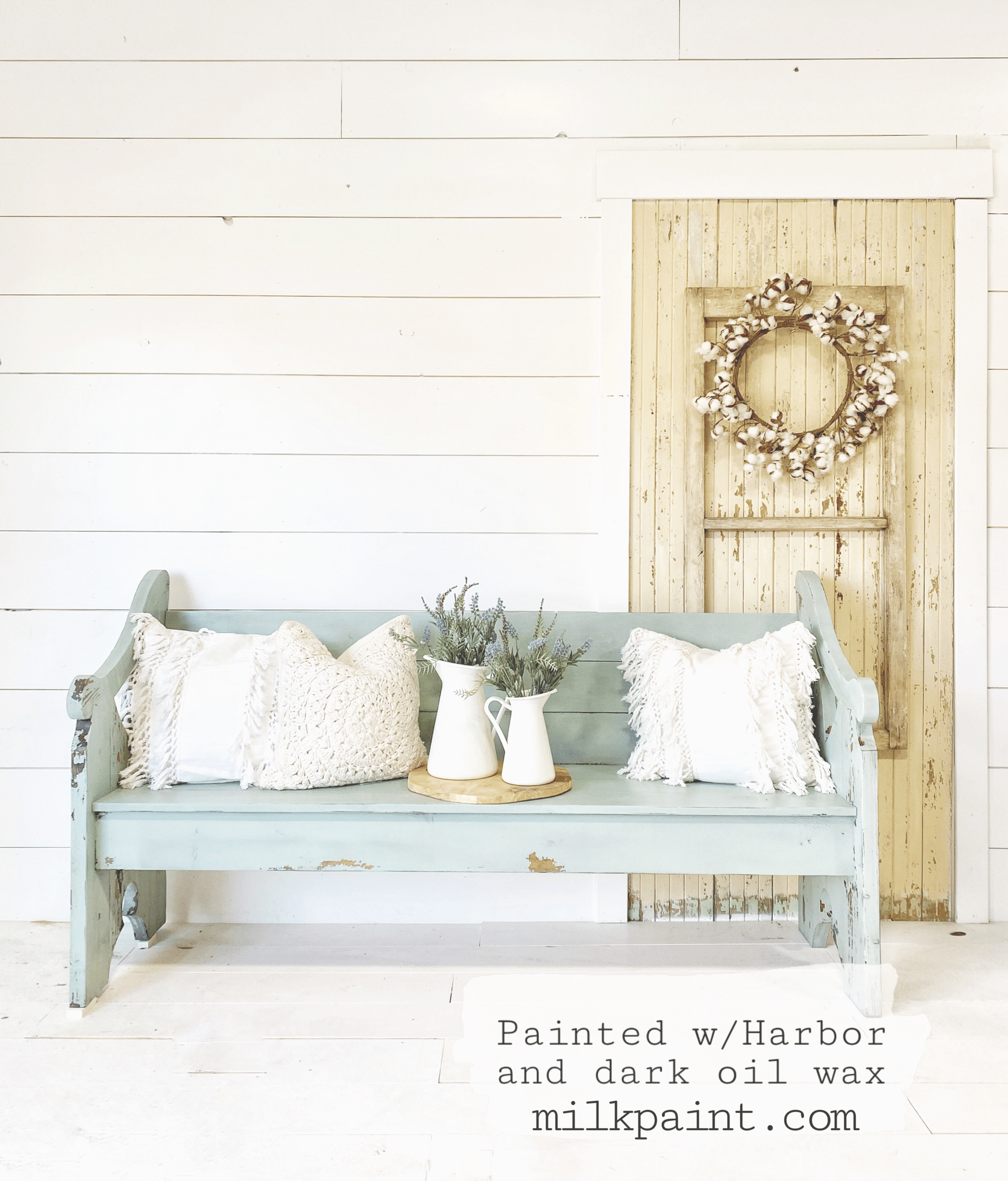 Light blue bench with decorative pillows and a wreath on a white wall background