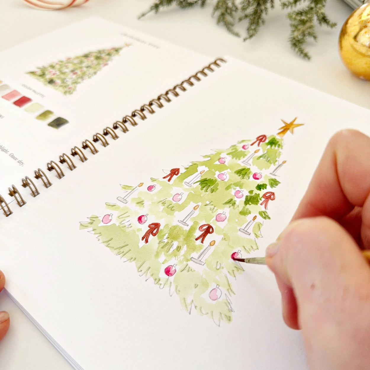 Person watercolor painting a Christmas tree on a spiral-bound notebook with a pen.