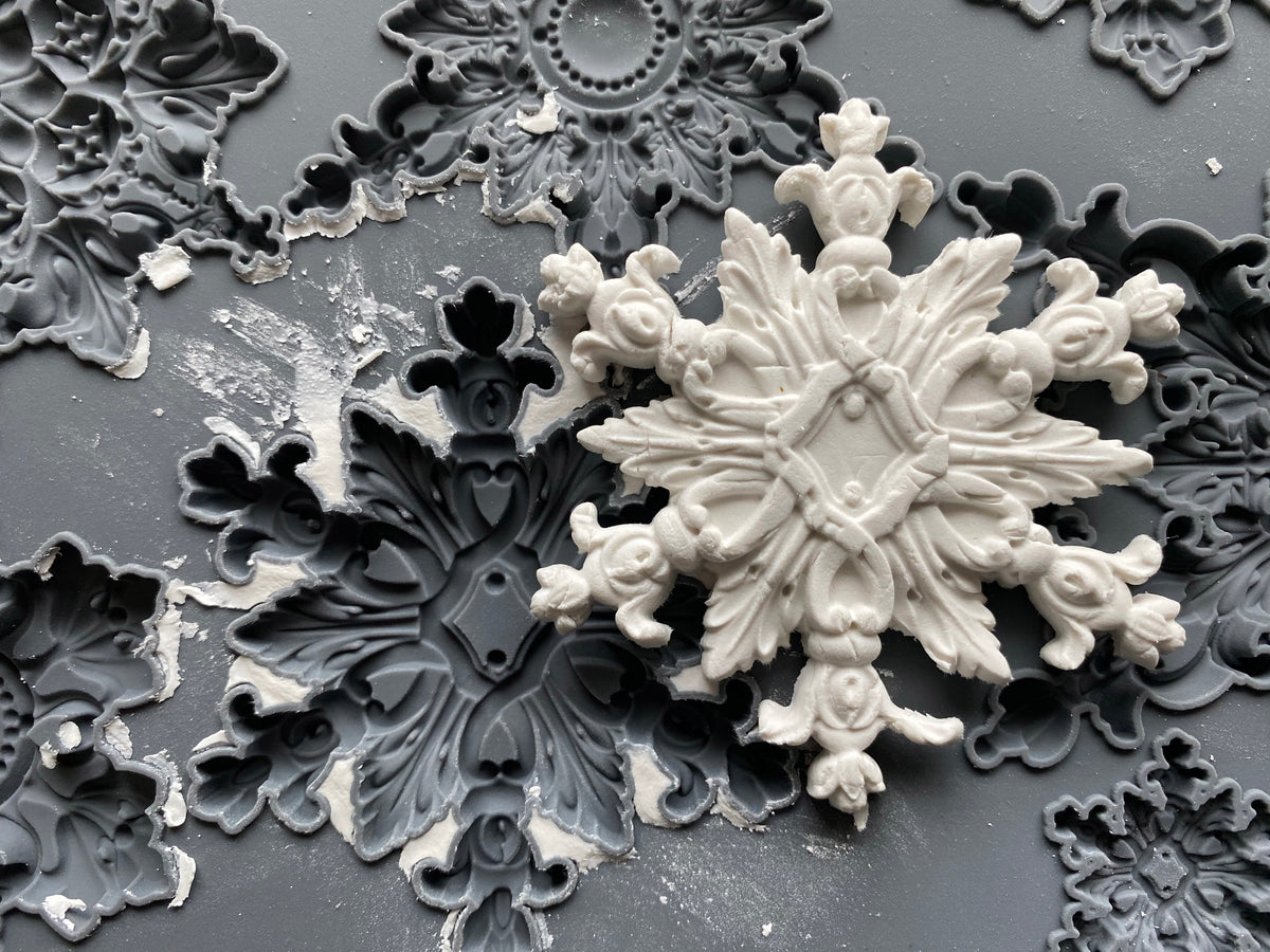 A close-up of a white ornate snowflake-shaped decorative object resting on a dark gsurface with matching gray ornamental molds or trays.