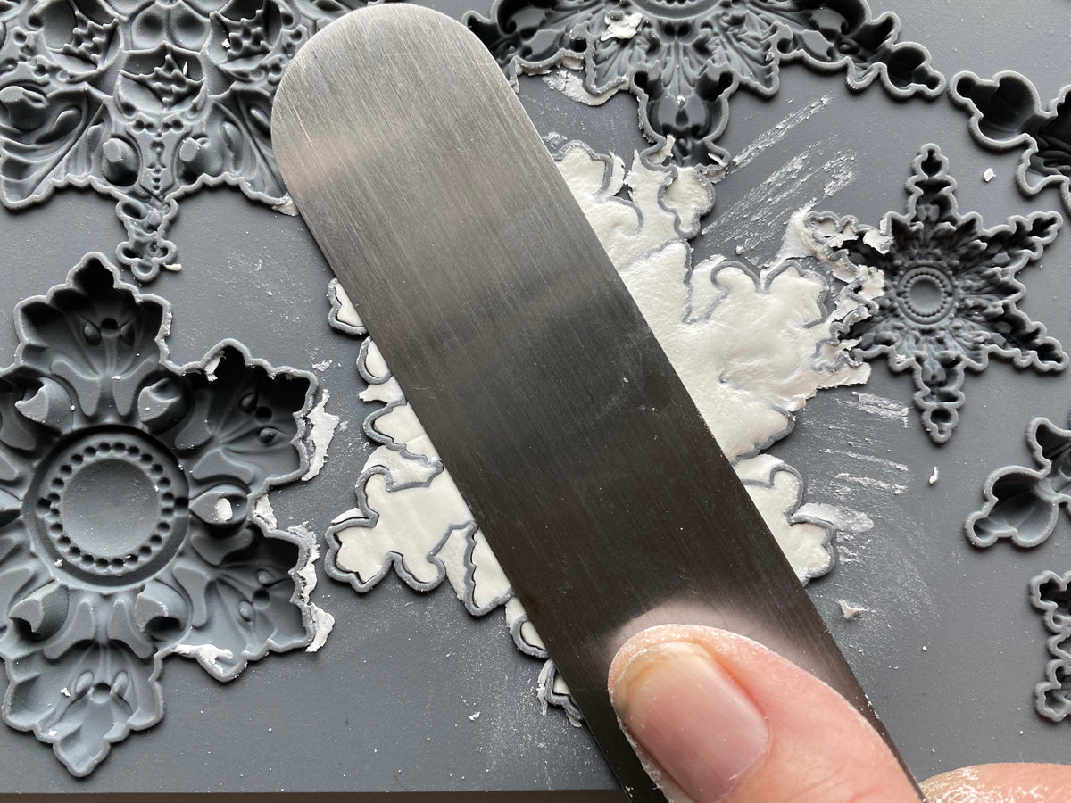 A close-up of a hand using a flat metal spatula to spread air dry clay or putty over a silicone mould decorated with ornate snowflake and floral embossed designs.