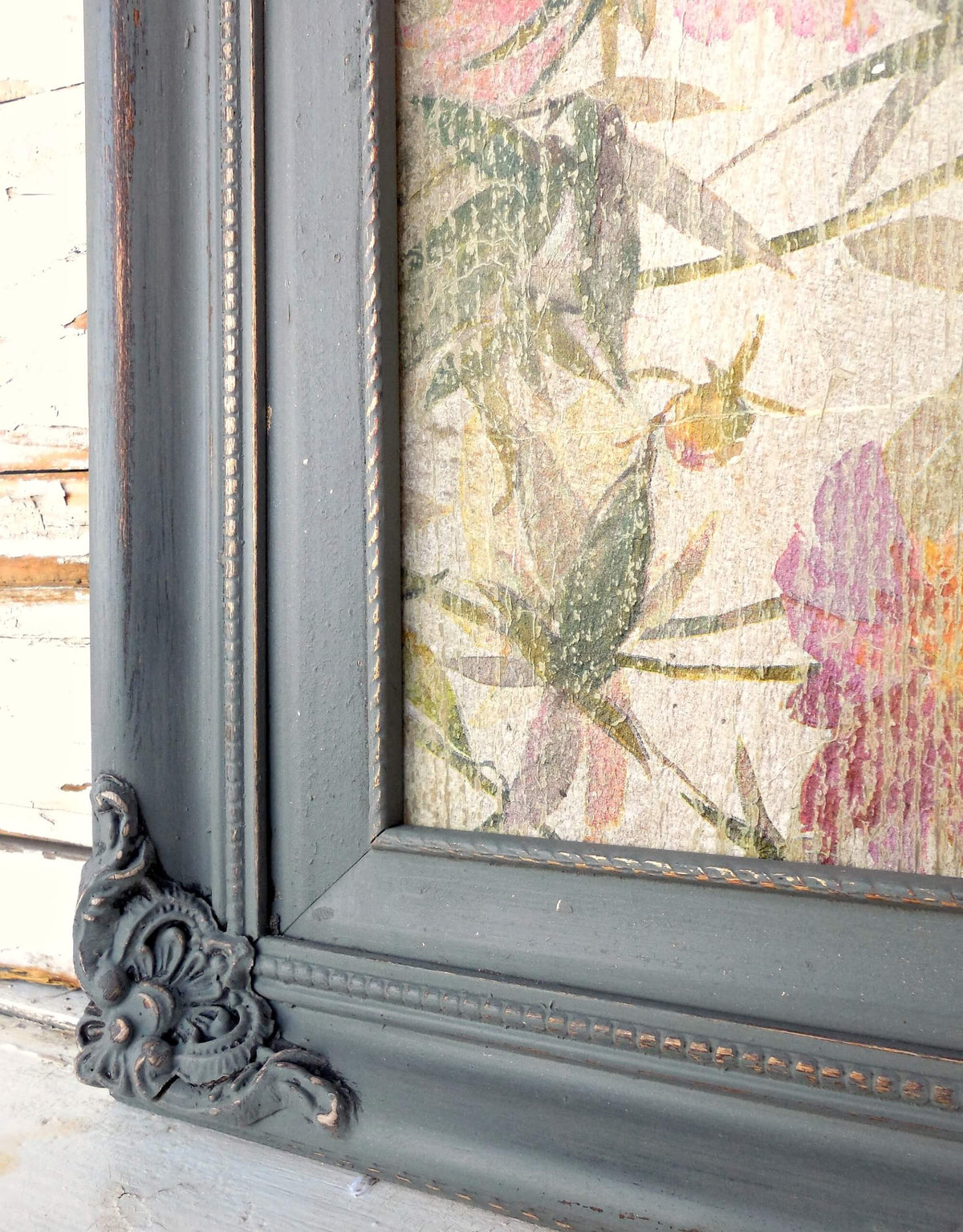 Floral artwork in a decorative grey milk-painted frame on a wood surface.