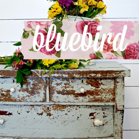 A weathered wooden drawer with the text 'bluebird' overlaid on top, surrounded by a floral pattern, suggesting the color of the milk paint.