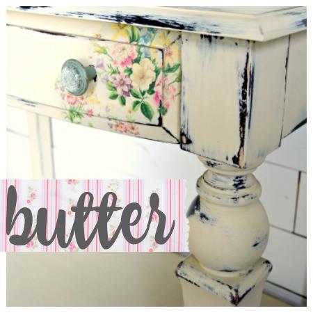 A close-up of a wooden furniture piece painted with Sweet Pickins Milk Paint in a white color, designated as 'butter'. The paint is applied in a distressed manner, showing the underlying floral patterned wallpaper.