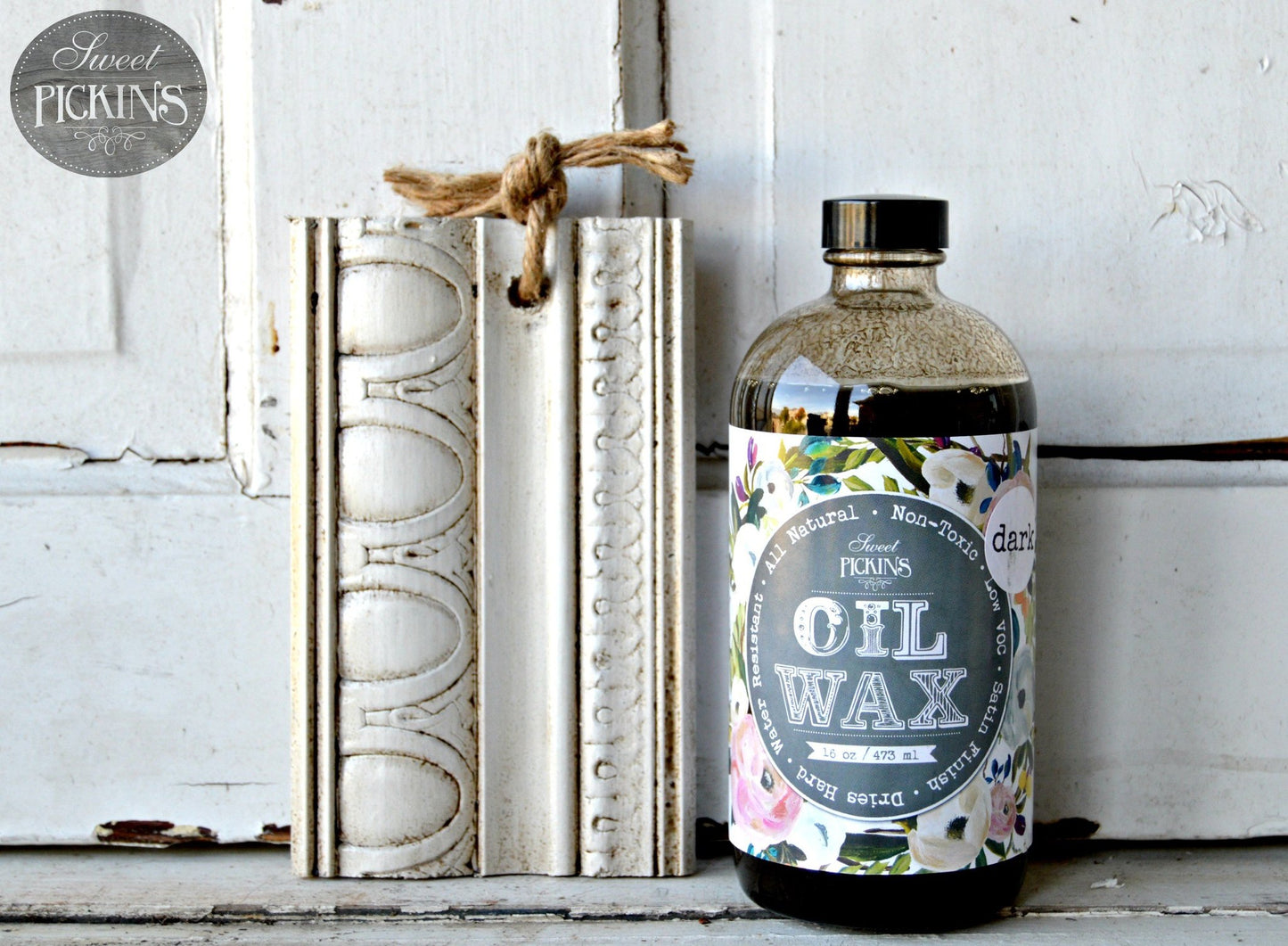 A bottle of Sweet Pickins Oil Wax with a dark tint, placed next to a decorated white frame on a wooden surface.