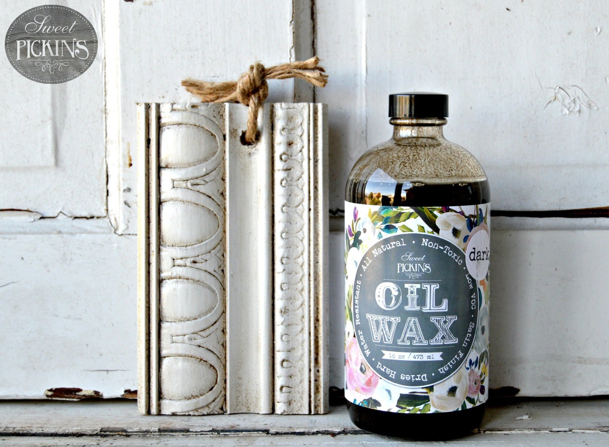 A bottle of Sweet Pickins Oil Wax with a dark tint, placed next to a decorated white frame on a wooden surface.