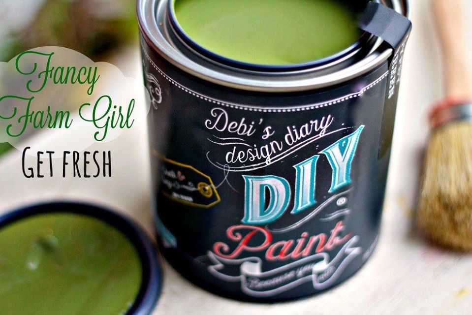 A can of 'Fancy Farm Girl' green paint from the brand DIY Paint Co. with a brush and a lid in the background.