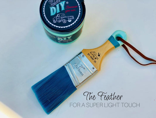 A brush from "DIY Paint Co." called the "Feather" brush lying on a table next to DIY Paint Co. brand paint.